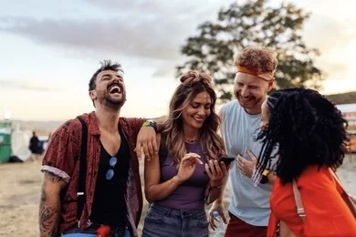 four friends laughing together outdoors at golden hour, one holding a phone